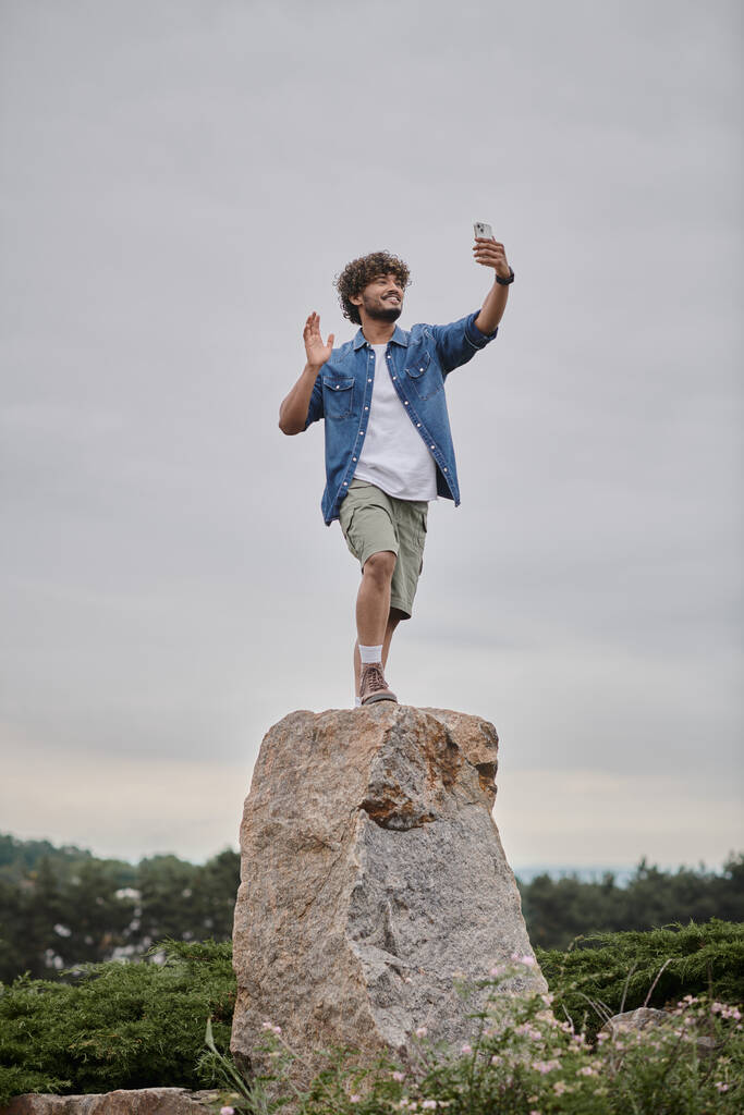concetto nomadismo, uomo indiano riccio in piedi sulla roccia e avendo video chat su smartphone, mano d'onda - Foto, immagini