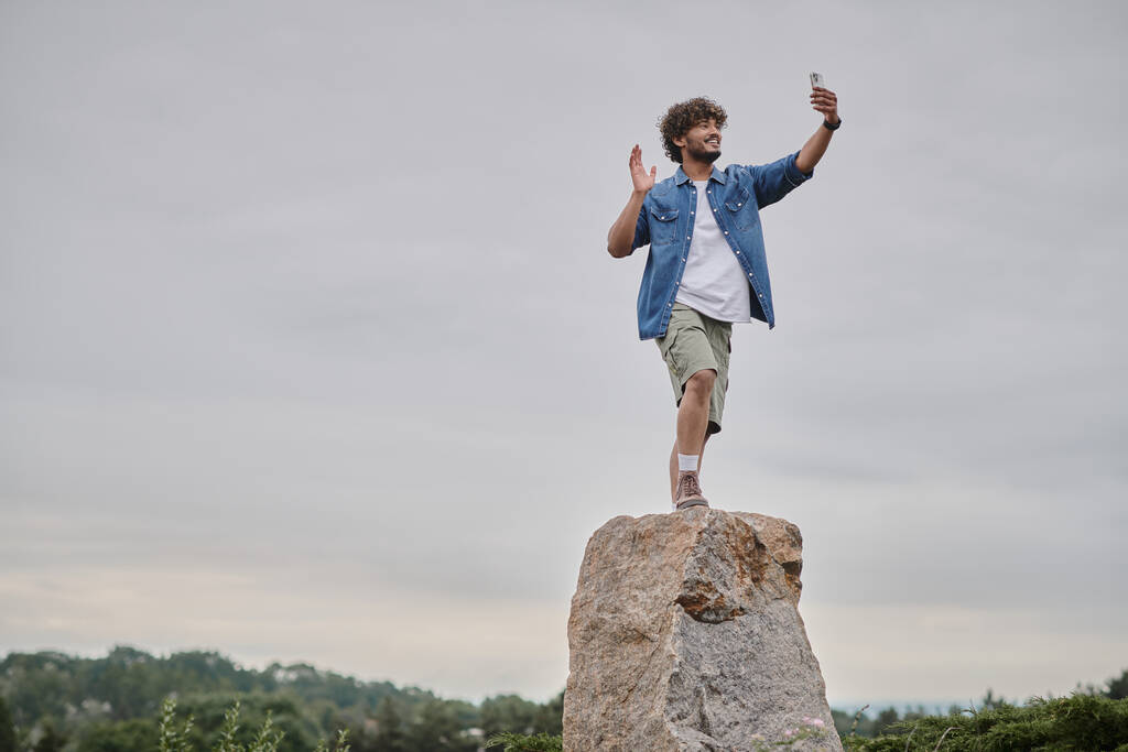 νομαδισμός έννοια, ευτυχισμένος ινδός άνθρωπος στέκεται στο βράχο και να έχουν συνομιλία βίντεο στο smartphone, χέρι κύμα - Φωτογραφία, εικόνα