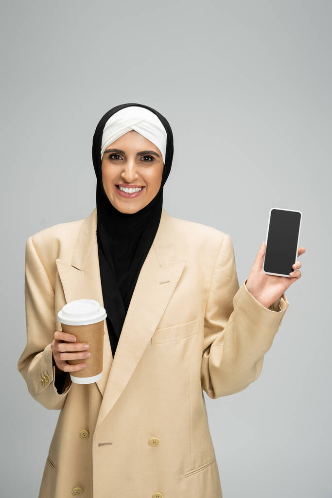 Gülümseyen, modaya uyan Müslüman iş kadını elinde kahve ile gri ekranda boş ekranlı akıllı telefonu gösterecek. - Fotoğraf, Görsel