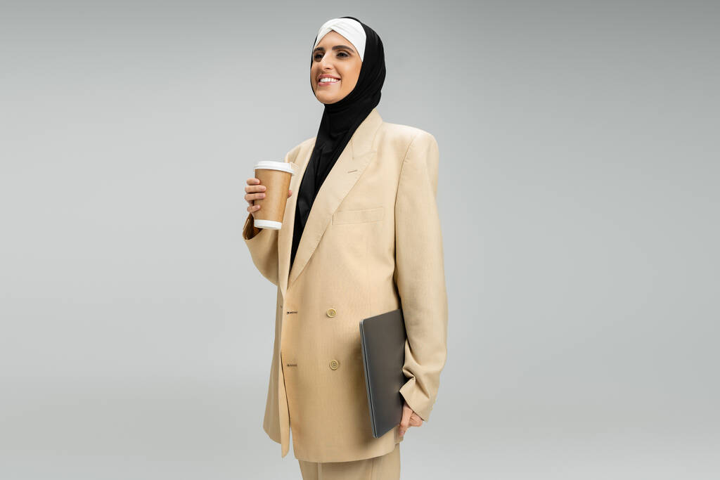 progresista y elegante mujer de negocios musulmana con bebida para llevar y portátil sonriendo en gris - Foto, Imagen