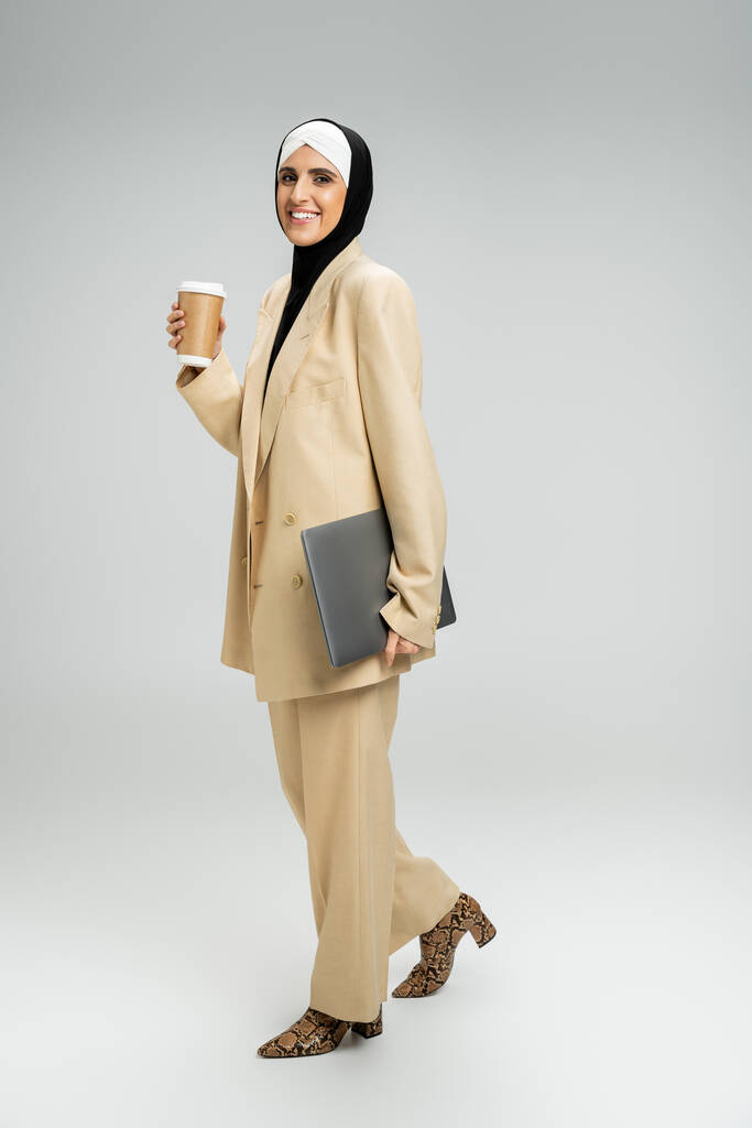 onnistunut musliminainen liikenainen, jolla on paperimuki, kannettava tietokone ja säteilevä hymy harmaalla, täyspitkä - Valokuva, kuva