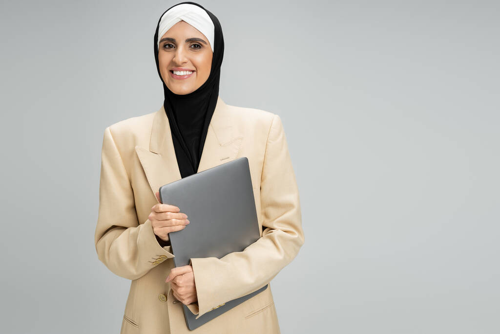 headshot of smiling muslim businesswoman in blazer and hijab holding laptop on grey, success - Photo, Image