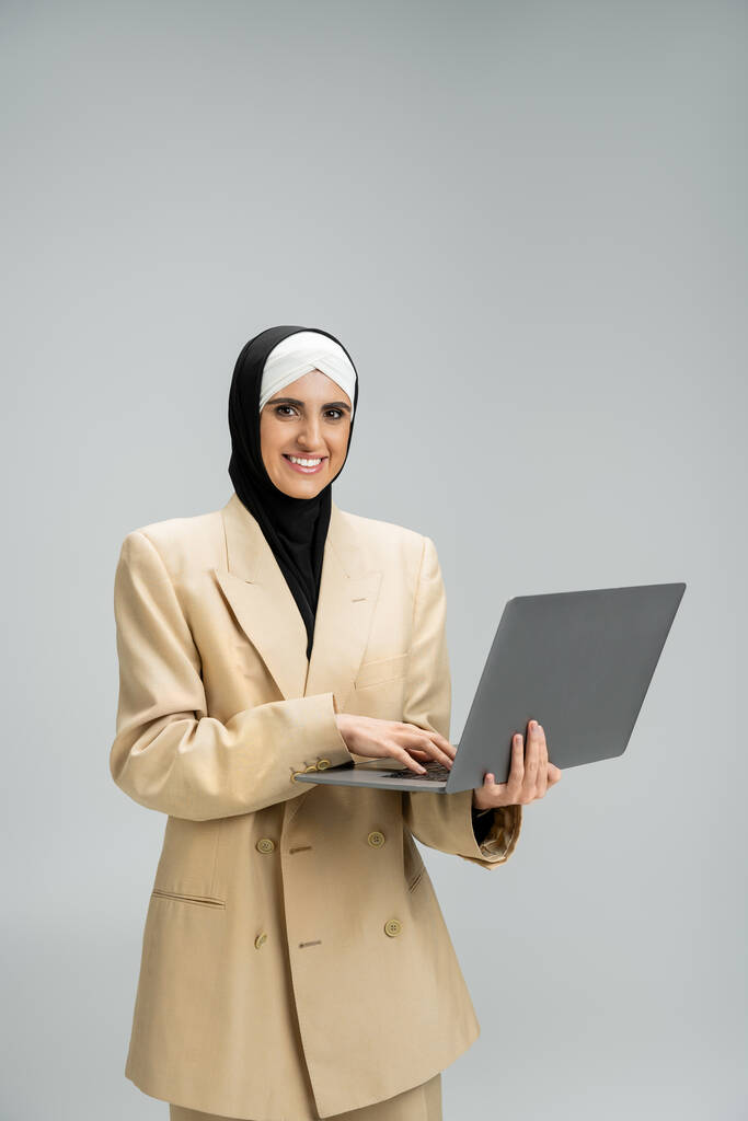 exitosa mujer de negocios musulmana en hijab y blazer celebración de la computadora portátil y sonriendo a la cámara en gris - Foto, imagen