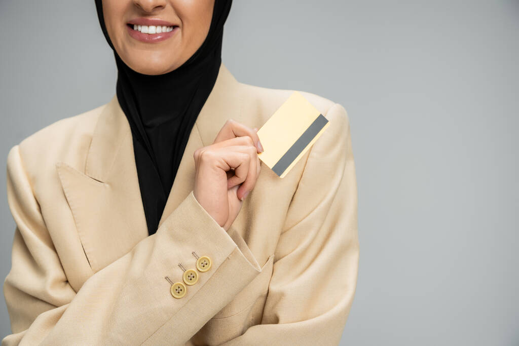 обрезанный вид улыбающейся мусульманской предпринимательницы в блейзере и хиджабе, держащей кредитную карту на серой - Фото, изображение