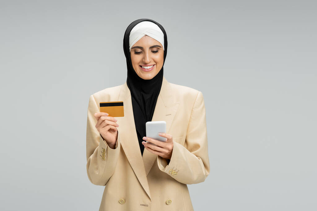 mujer de negocios musulmana complacida en hijab y elegante chaqueta con teléfono inteligente y tarjeta de crédito en gris - Foto, Imagen