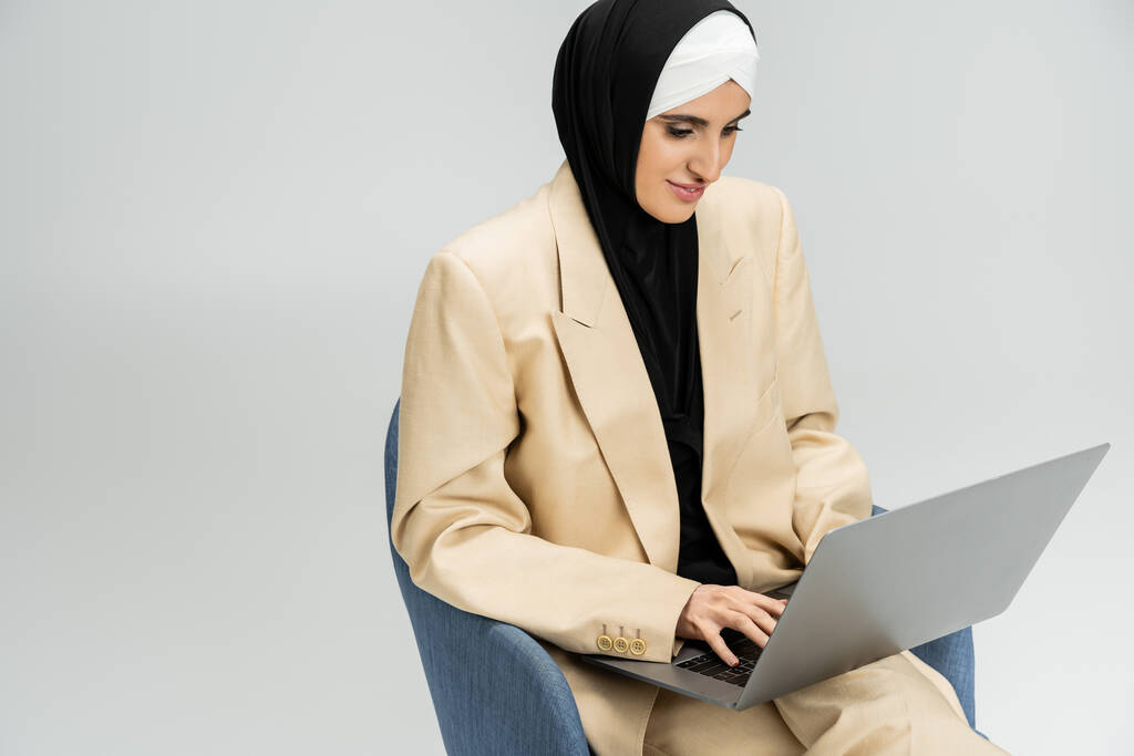 συμπυκνωμένο muslim επιχειρηματίας με κοστούμι και hijab δακτυλογράφηση σε φορητό υπολογιστή σε πολυθρόνα σε γκρι - Φωτογραφία, εικόνα