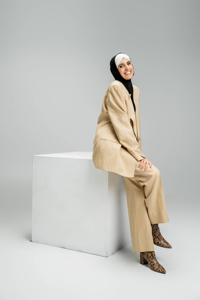 joyful muslim businesswoman in hijab and beige suit sitting on white cube on grey, full length - Photo, Image