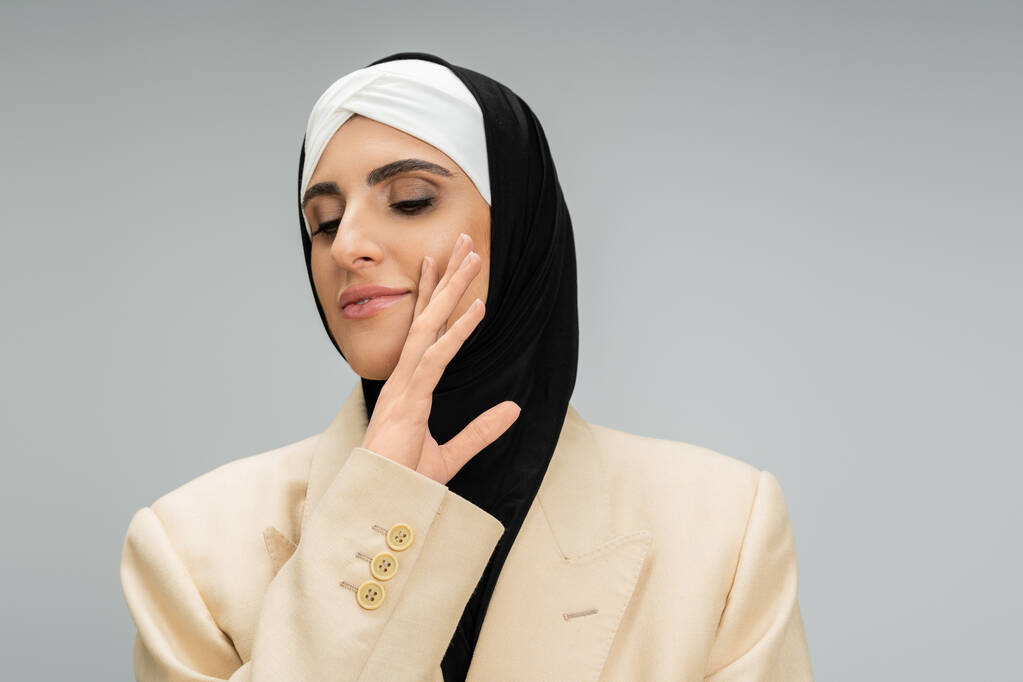 expressive, stylish muslim businesswoman with closed eyes and makeup holding hand near chin on grey - Photo, Image
