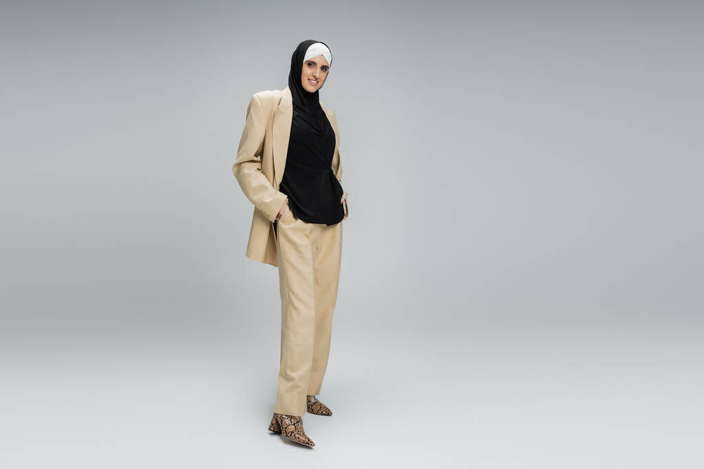 cheerful muslim businesswoman in beige trendy suit and hijab posing with hands in pockets on grey - Photo, Image