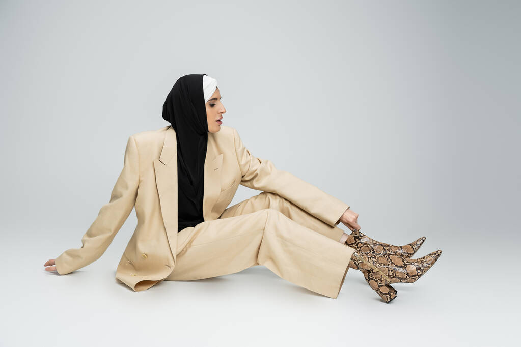 модна мусульманська бізнес-леді в хіджабі та бежевому костюмі, що регулює завантаження змій на сірому - Фото, зображення