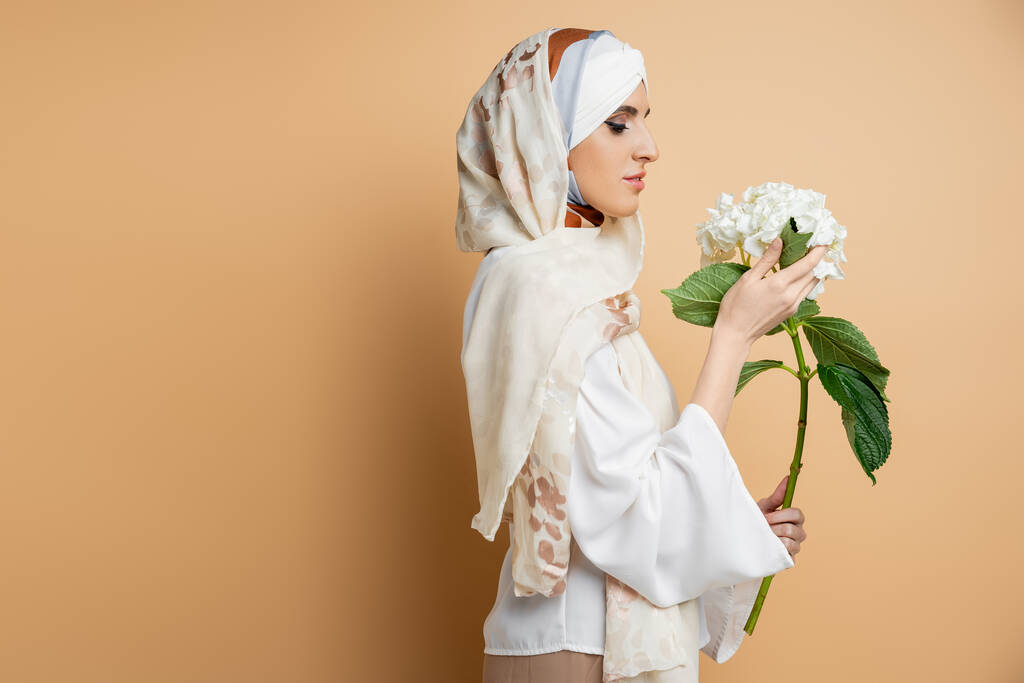 mujer musulmana elegante en pañuelo de seda y blusa sosteniendo flor blanca en beige, vista lateral - Foto, Imagen