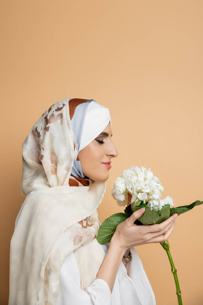 ベージュ,サイドビューに白いハイドレンジアの花を保持するエレガントなヘッドスカーフで喜んだイスラム教徒の女性 - 写真・画像