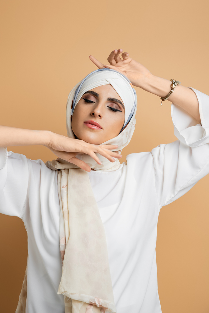 mujer musulmana sensual con los ojos cerrados y maquillaje posando en pañuelo de seda y blusa blanca en beige - Foto, Imagen