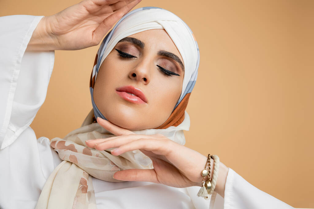 όμορφη μουσουλμάνα γυναίκα με τα χέρια κοντά στο πρόσωπο και κλειστά μάτια σε μεταξωτή μαντίλα σε μπεζ, πορτρέτο - Φωτογραφία, εικόνα
