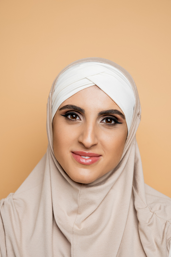 πορτρέτο χαρούμενη μουσουλμάνα γυναίκα με μακιγιάζ φορώντας μαντίλα και κοιτάζοντας κάμερα σε μπεζ - Φωτογραφία, εικόνα