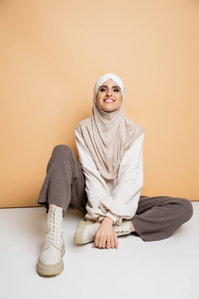 ξέγνοιαστη μουσουλμάνα γυναίκα σε μοντέρνα casual στολή και μαντίλα κάθεται σε μπεζ, σύγχρονο τρόπο ζωής - Φωτογραφία, εικόνα