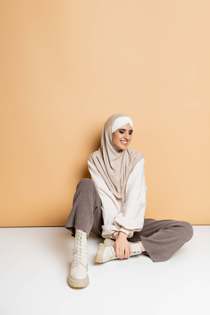 beztroska muzułmanka w modnym stroju casual i hidżab siedzi na beżowym, pełnej długości - Zdjęcie, obraz