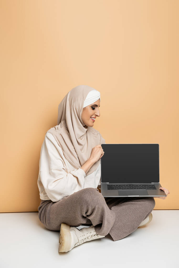 happy muslim woman in hijab and stylish casual attire sitting with laptop with blank screen on beige - Photo, Image