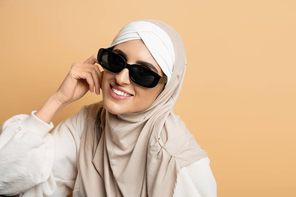tesettür ve güneş gözlüklü neşeli ve moda müslüman kadın kamera bej, modern yaşam tarzı gülümsüyor - Fotoğraf, Görsel