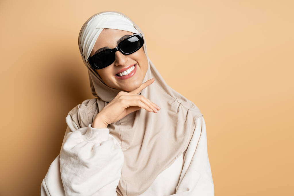 χαρούμενη μουσουλμάνα γυναίκα με μαντίλα και μοντέρνα γυαλιά ηλίου ποζάροντας με το χέρι κοντά στο πηγούνι και χαμογελώντας σε μπεζ - Φωτογραφία, εικόνα