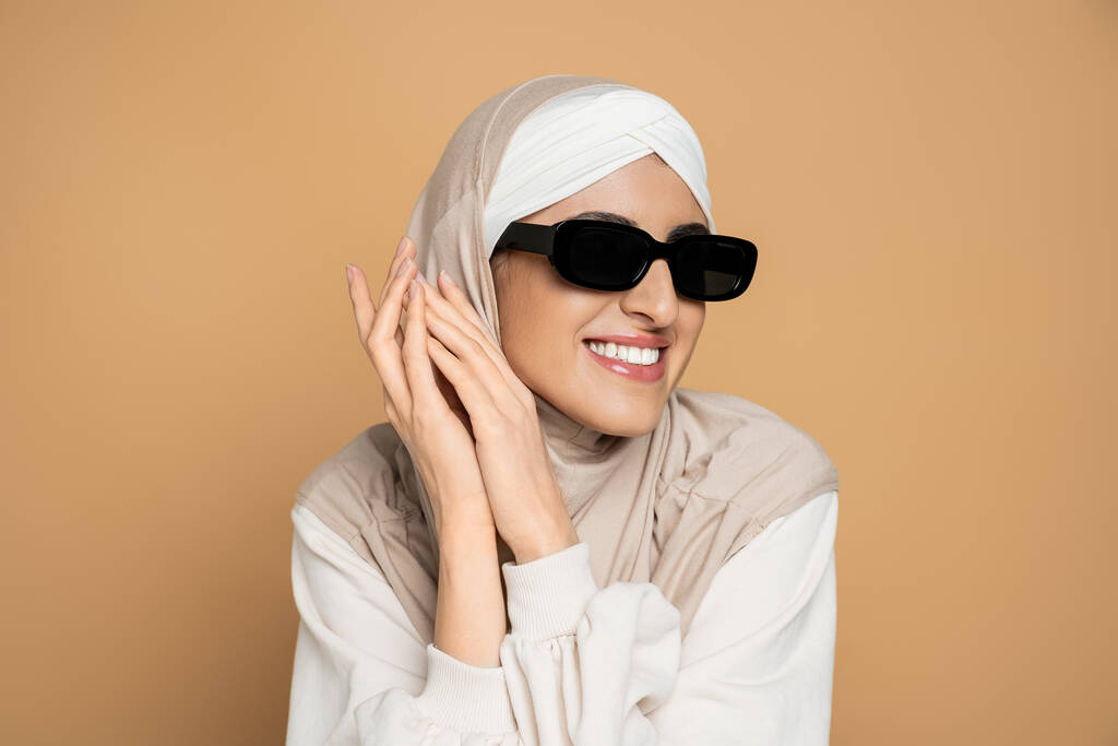 mujer musulmana alegre en hijab, gafas de sol de moda y sudadera blanca, con las manos cerca de la cara en beige - Foto, Imagen