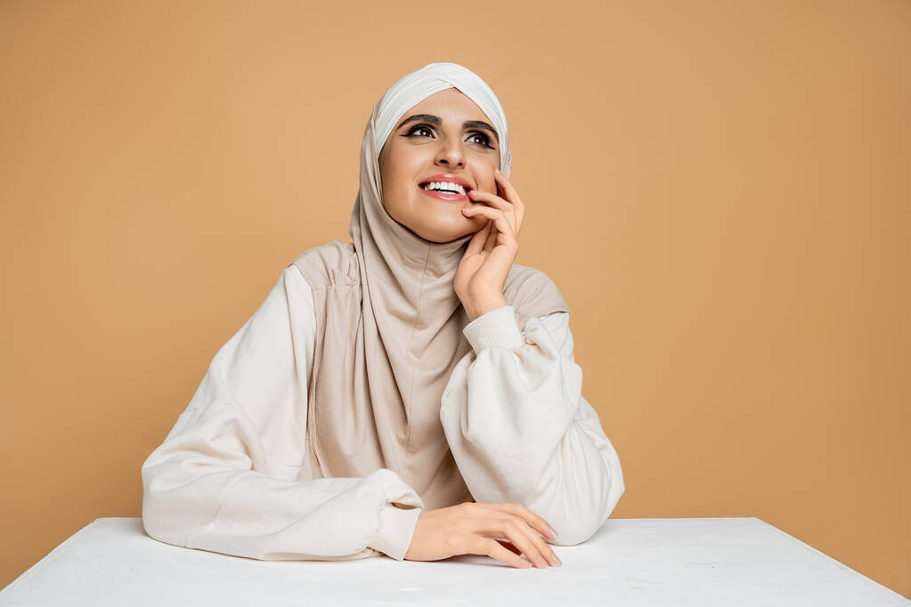 Modern Müslüman kadın tesettürlü ve sweatshirt 'lü beyaz masada oturuyor ve bej renge bakıyor. - Fotoğraf, Görsel
