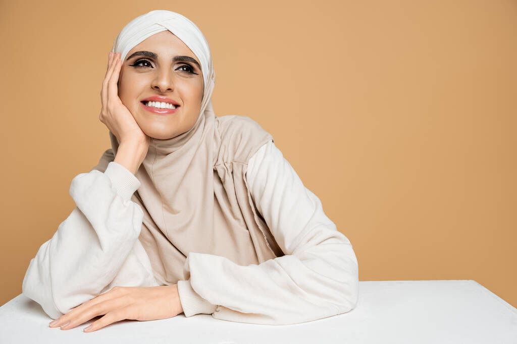 gelukkig en dromerig moslim vrouw in stijlvolle sweatshirt en hijab weg te kijken aan witte tafel op beige - Foto, afbeelding