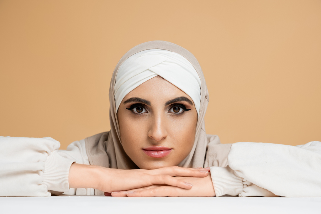 attraktive muslimische Frau mit Make-up, die im Hijab am weißen Tisch sitzt und in beige in die Kamera schaut - Foto, Bild