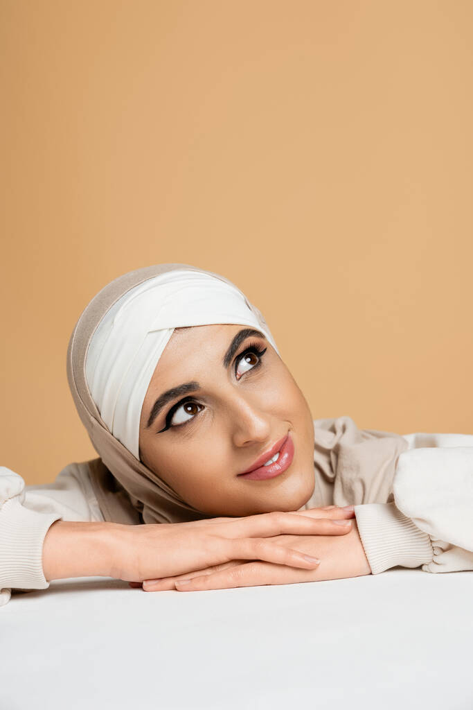álmodozó muszlim nő hidzsábban mosolyog, és elfordítja tekintetét asztalra bézs, kifejező egyéniség - Fotó, kép