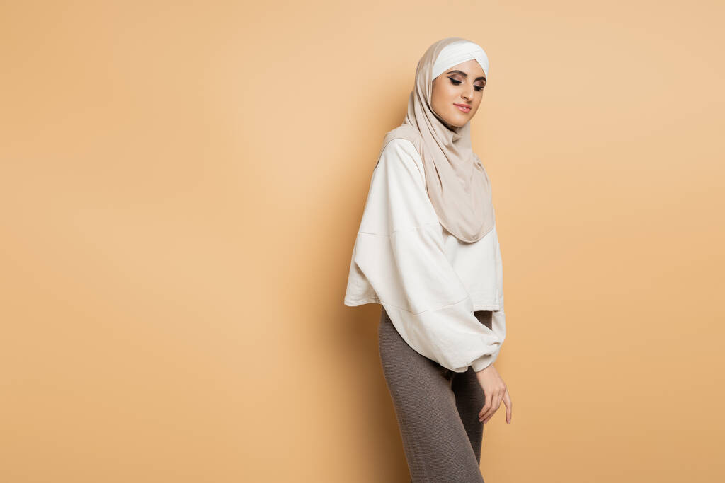 czarująca muzułmanka w hidżabie, biała bluza i szare spodnie stojące na beżowym, osobistym stylu - Zdjęcie, obraz