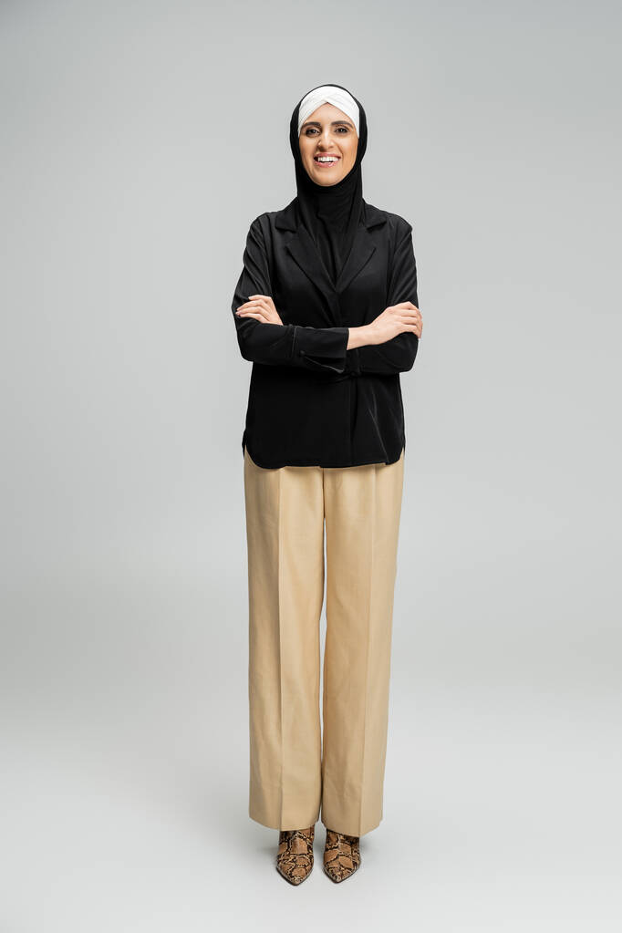 счастливая мусульманка-бизнесвумен в хиджабе и стильном наряде, позирующая со сложенными руками на серой, полной длине - Фото, изображение