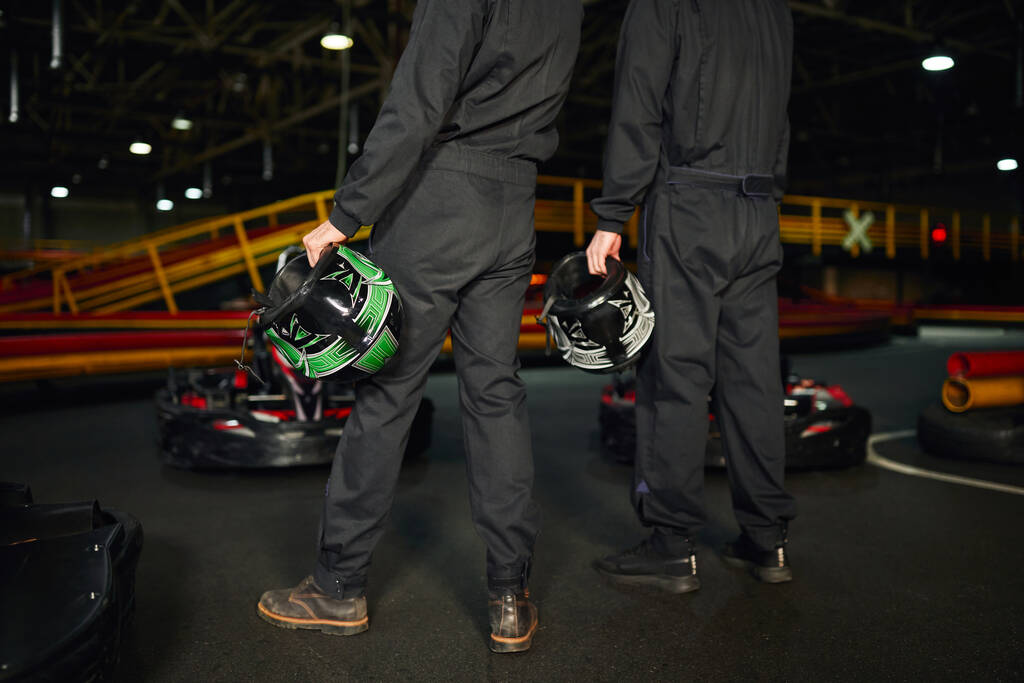vista recortada de dos hombres de pie cerca de los coches de carreras y la celebración de cascos, el equipo de conductores de karts - Foto, imagen