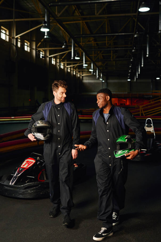 coureurs de kart interracial marchant près des voitures de course et tenant des casques, les hommes à l'intérieur du circuit de kart - Photo, image