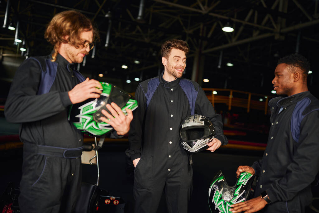позитивные мультикультурные мужчины болтают внутри картинговой цепи, три гонщика на картинге держат шлемы - Фото, изображение