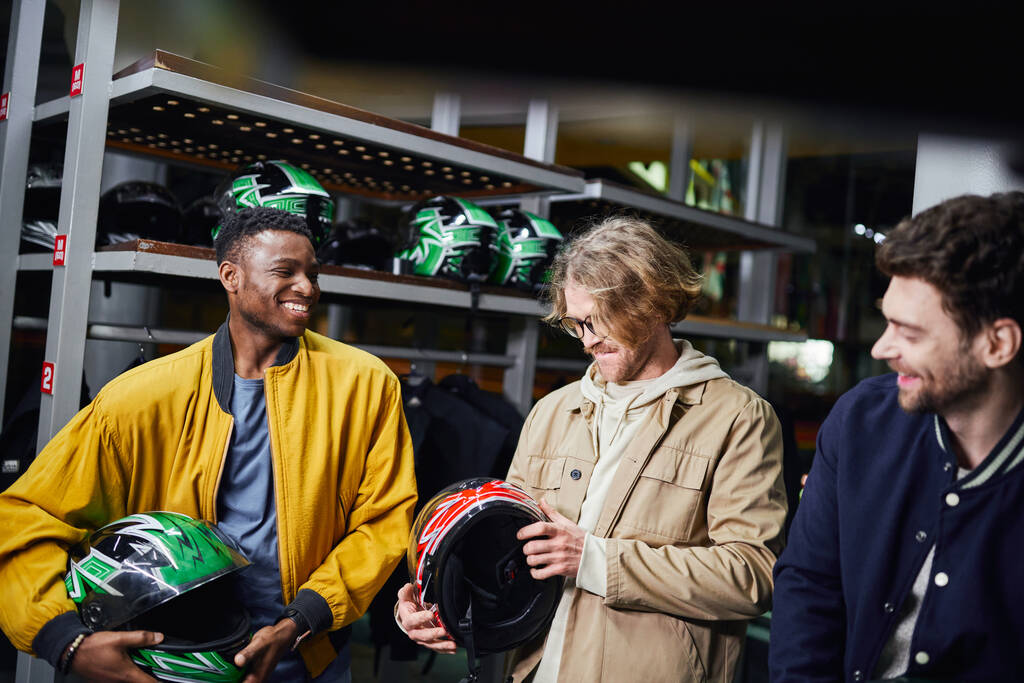 fröhliche gemischtrassige männliche Freunde in Jacken mit Helmen, Indoor-Rennbahn, Kart-Konzept - Foto, Bild