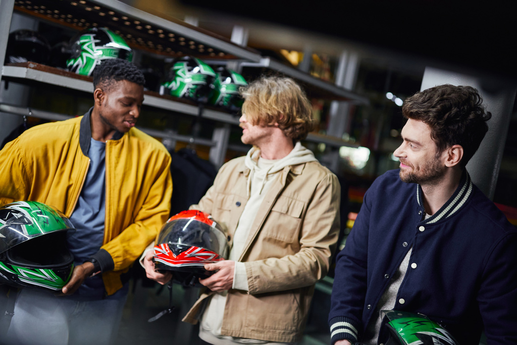 felizes amigos masculinos multiculturais em jaquetas segurando capacetes, pista de corrida indoor, conceito de karting - Foto, Imagem