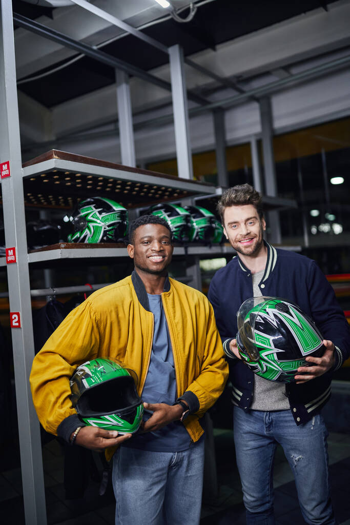 два мультикультурных друга, держащих шлемы и смотрящих в камеру внутри картинговой дорожки, повозка - Фото, изображение