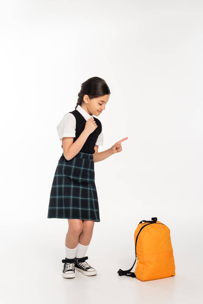 happy girl in school uniform standing and looking at backpack on white background, pointing away - Photo, Image