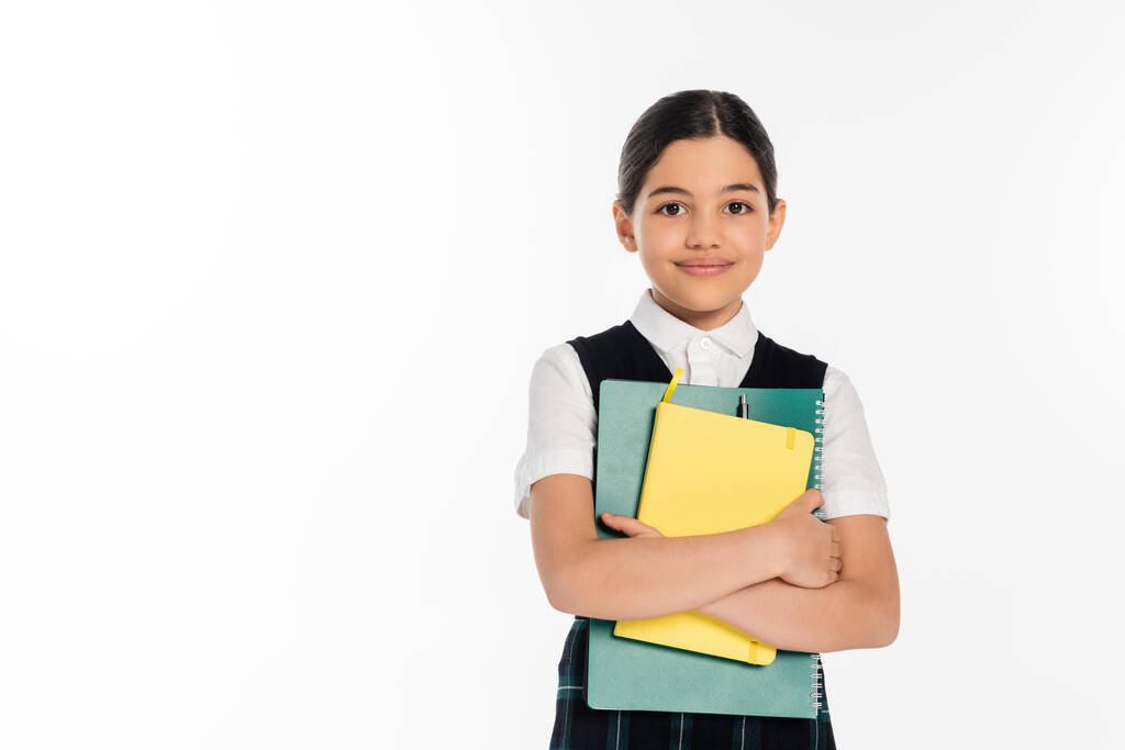 šťastná školačka stojící se zápisníky v rukou a dívající se na kameru izolovanou na bílém, studentka - Fotografie, Obrázek