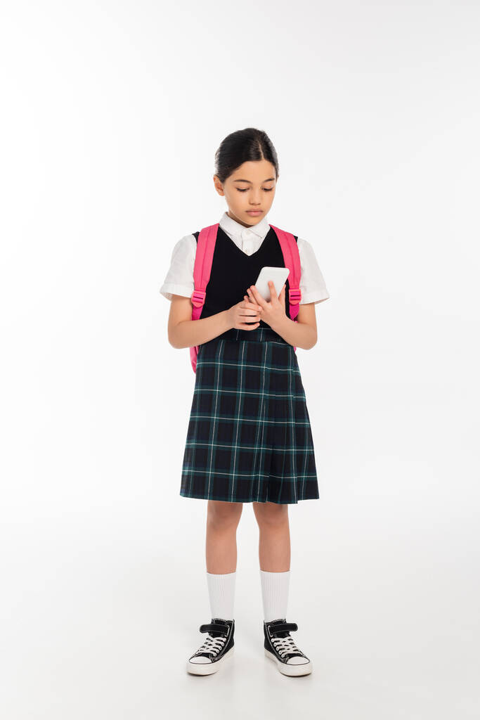digitální věk, školačka s batohem držící smartphone na bílém, studentka v uniformě, plná délka - Fotografie, Obrázek