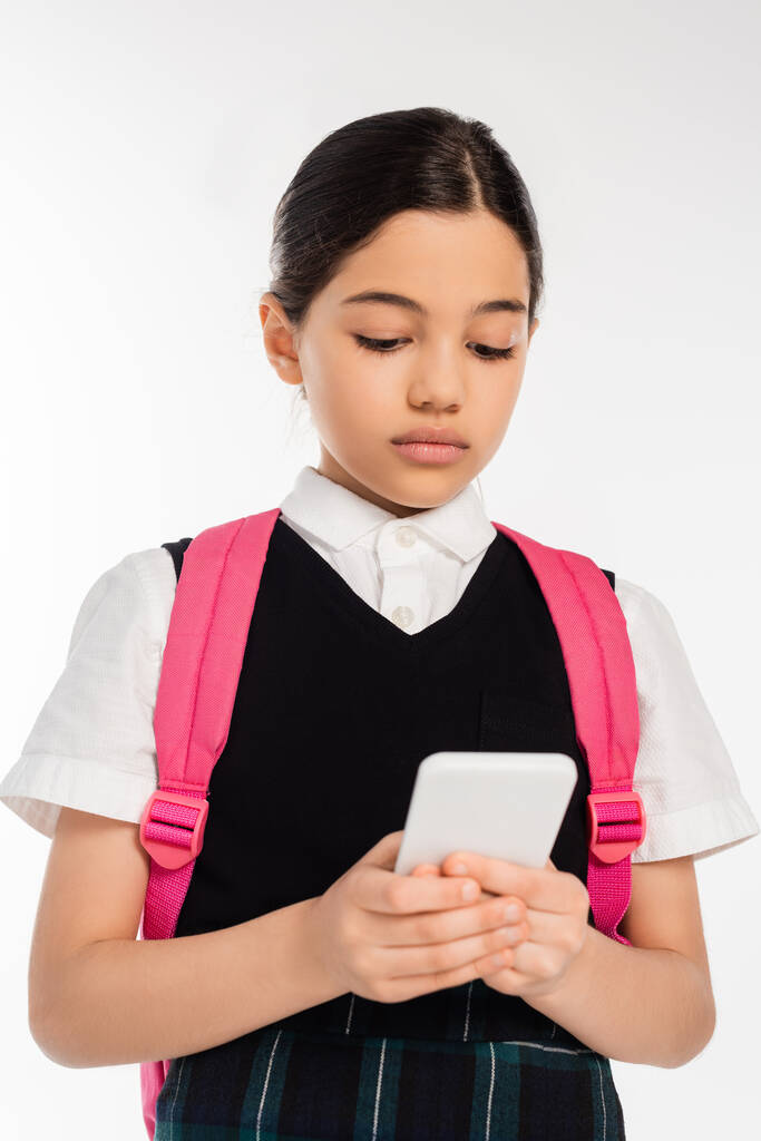âge numérique, écolière avec sac à dos en utilisant smartphone isolé sur blanc, étudiante en uniforme - Photo, image