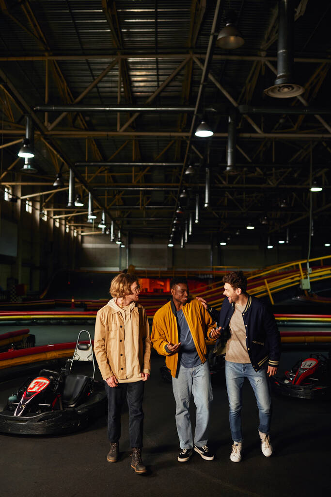 cheerful diverse friends chatting and walking near racing cars inside of karting track, hobby - Photo, Image