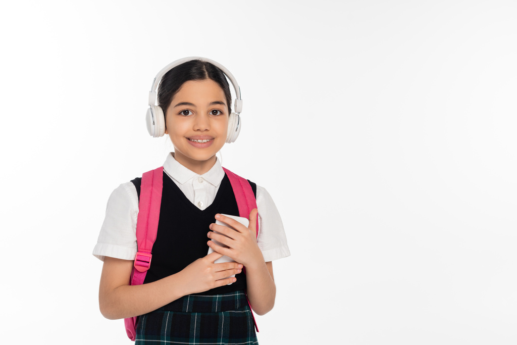 цифровой возраст, веселая школьница в беспроводных наушниках с помощью смартфона изолированы на белом, студент - Фото, изображение