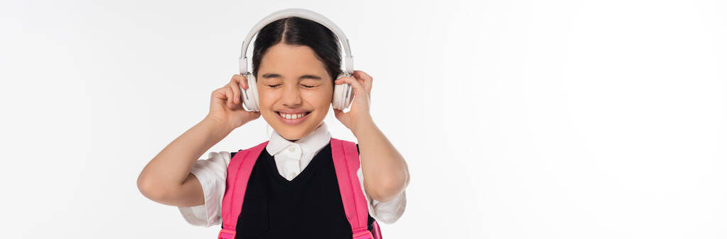 joyous schoolgirl in wireless headphones listening music isolated on white, closed eyes, banner - Photo, Image