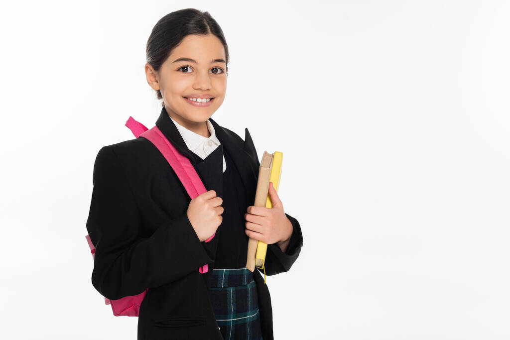 веселая школьница, стоящая с блокнотами и рюкзаком, изолированная на белом, возвращаясь к школьной концепции - Фото, изображение