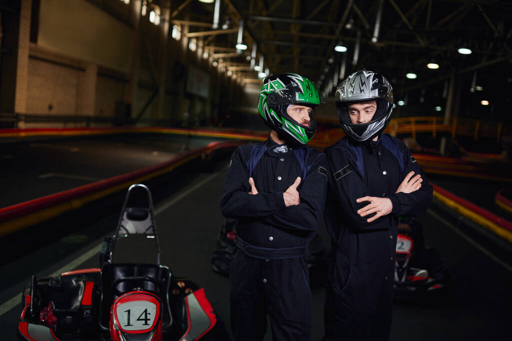 два уверенных в себе картинга гонщиков в спортивной одежде и шлемах, стоящих со сложенными руками на цепи - Фото, изображение