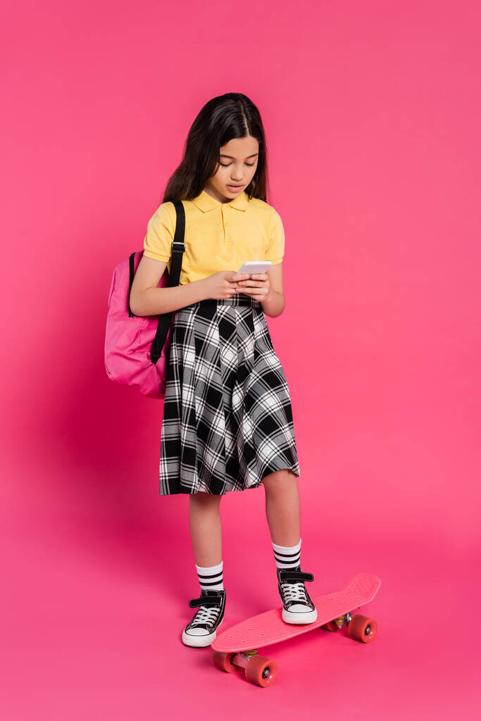 μελαχρινή μαθήτρια που χρησιμοποιεί smartphone και στέκεται κοντά στο penny board σε ροζ φόντο, νεανική ζωή - Φωτογραφία, εικόνα