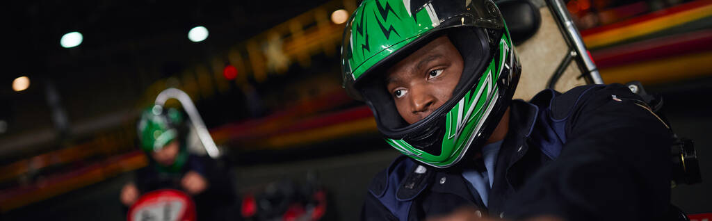 africano americano hombre en casco conducción ir kart en circuito cerca amigo en borrosa telón de fondo, bandera - Foto, imagen