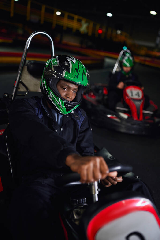Африканский американский водитель в шлеме вождение картинг на крытой трассе рядом друг на размытом фоне - Фото, изображение