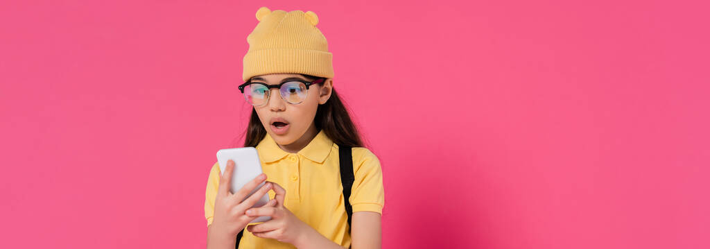 colegiala conmocionada en gorro sombrero y gafas con el teléfono inteligente sobre fondo rosa, elegante, pancarta - Foto, Imagen